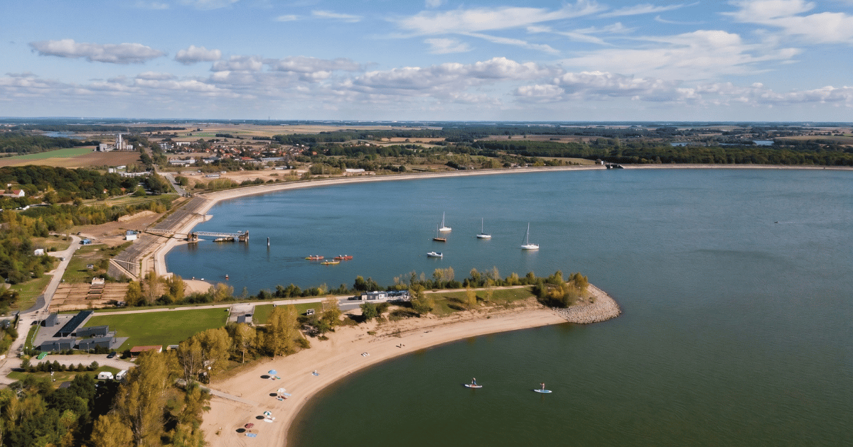 Widok z powietrza na Zalew Mietkowski: piaszczysta plaża z parasolami, paddleboardy i żaglówki na wodzie